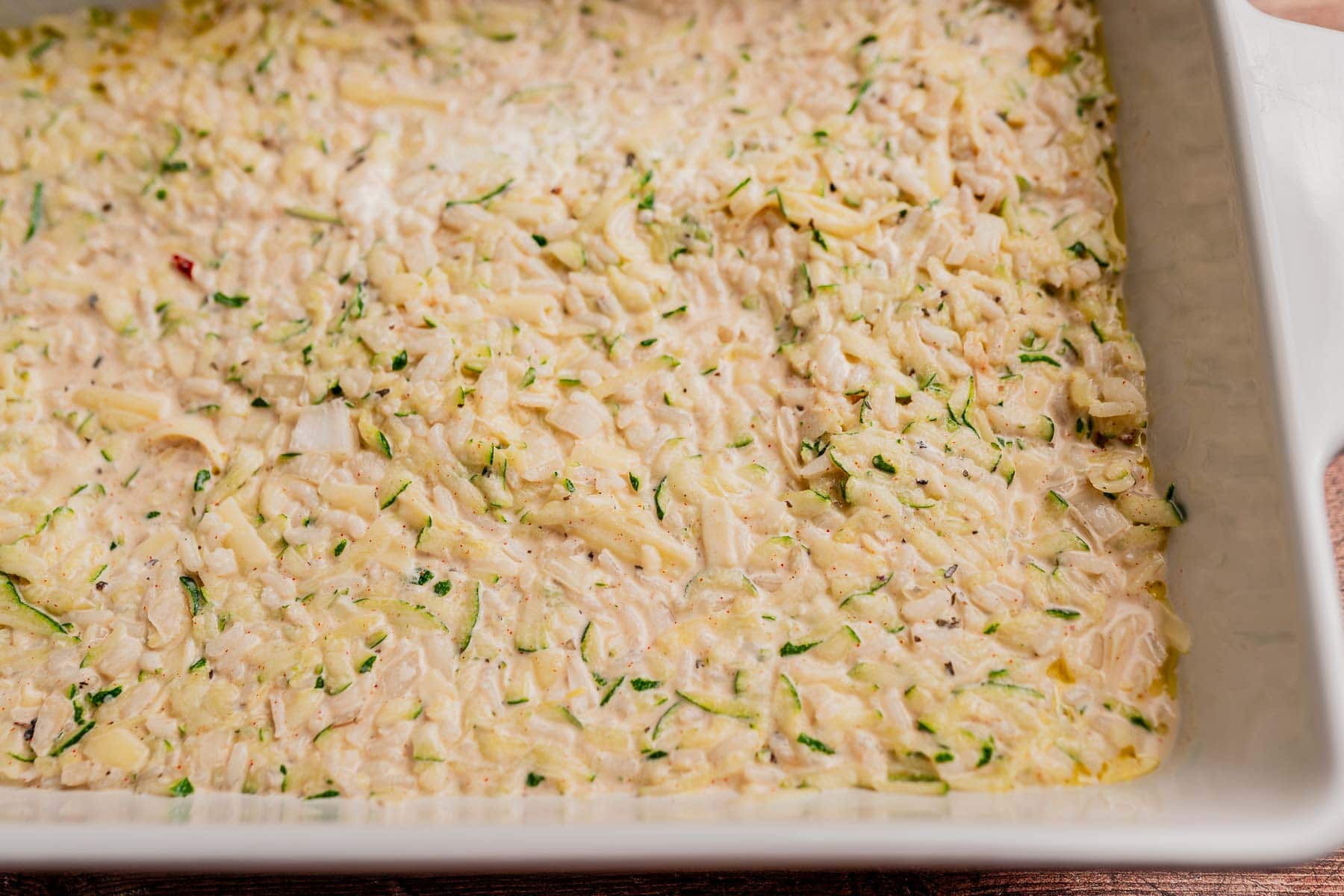 A close-up of an unbaked zucchini rice casserole mixture spread evenly in a rectangular baking dish.