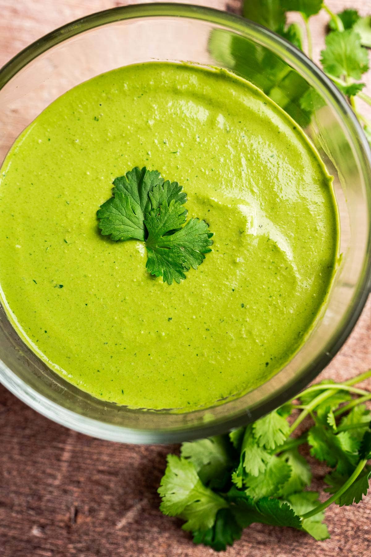 A glass bowl filled with creamy cilantro sauce, garnished with a cilantro leaf, sits on a wooden surface alongside fresh cilantro.