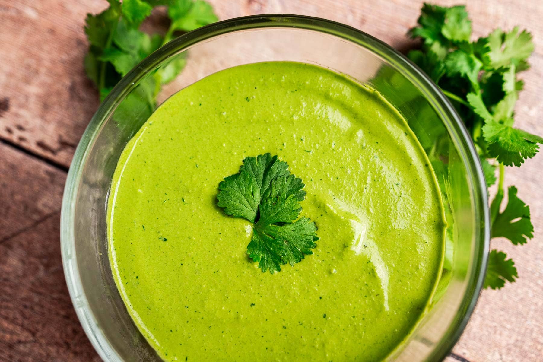 A glass bowl filled with creamy green cilantro sauce, topped with a sprig of cilantro, sits on a wooden surface with fresh cilantro leaves nearby.