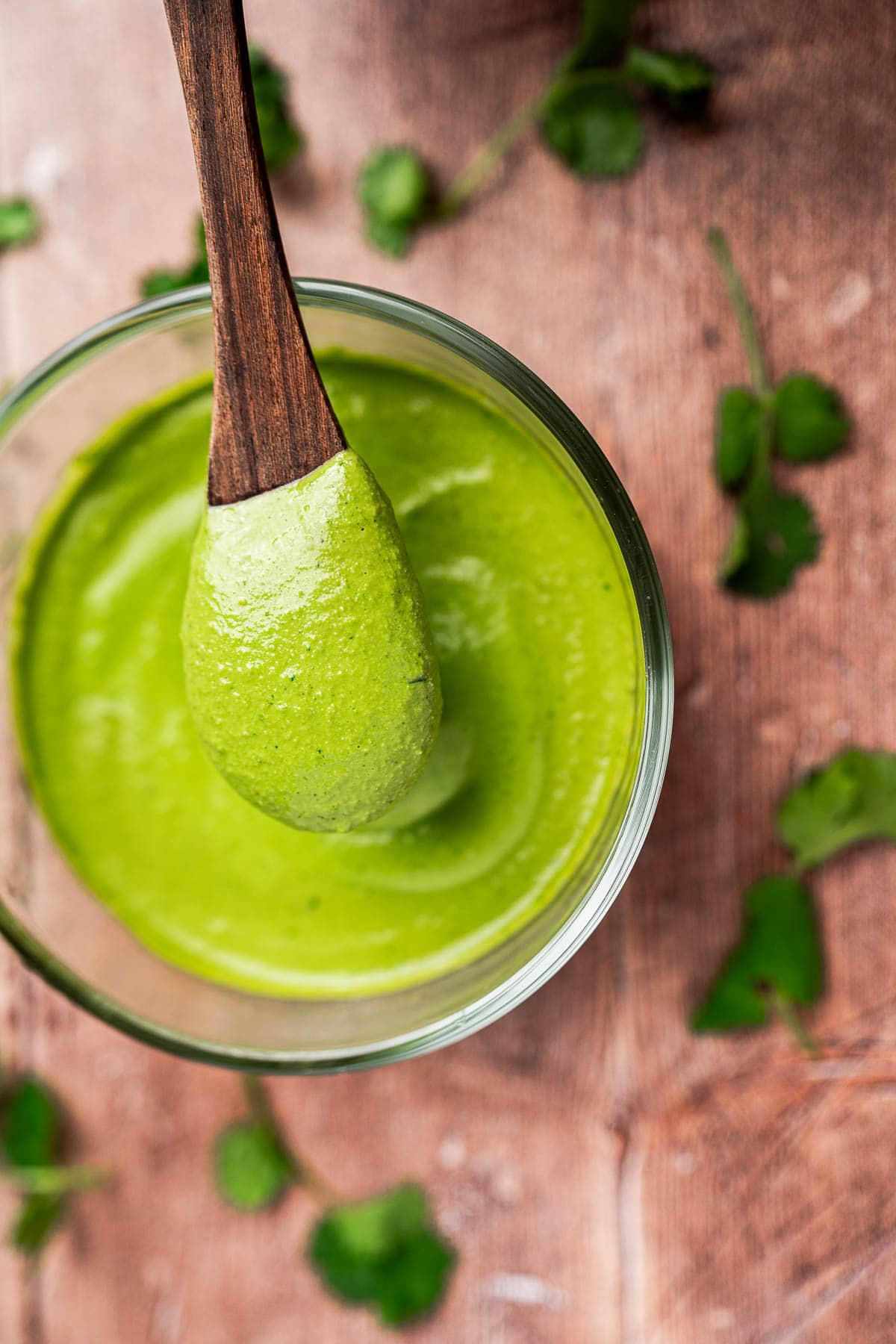 A wooden spoon holding cilantro sauce hovers over a glass bowl, with fresh cilantro leaves scattered across a wooden surface.