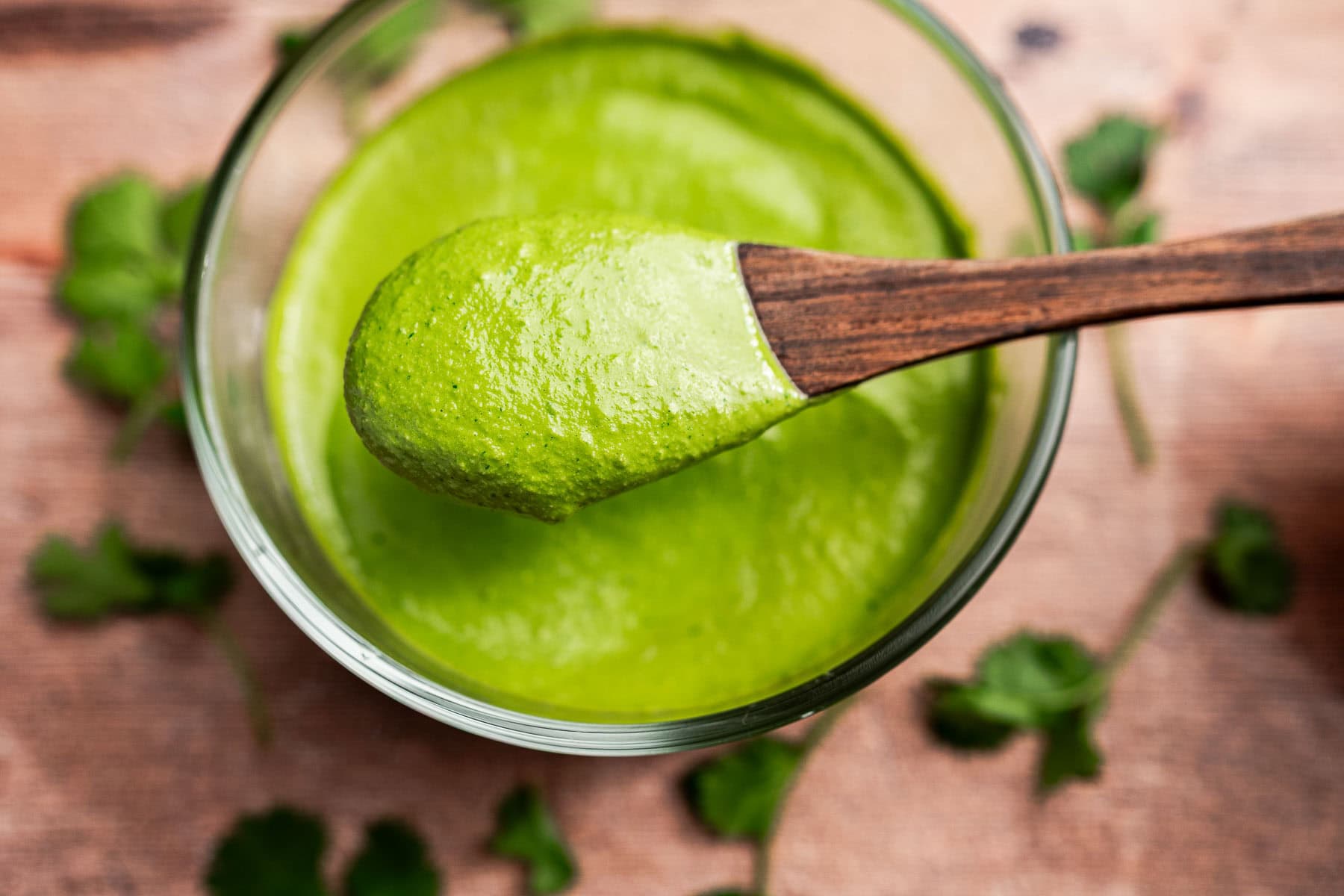 A wooden spoon holds a portion of thick green cilantro sauce above a glass bowl, with fresh cilantro leaves scattered on a wooden surface.