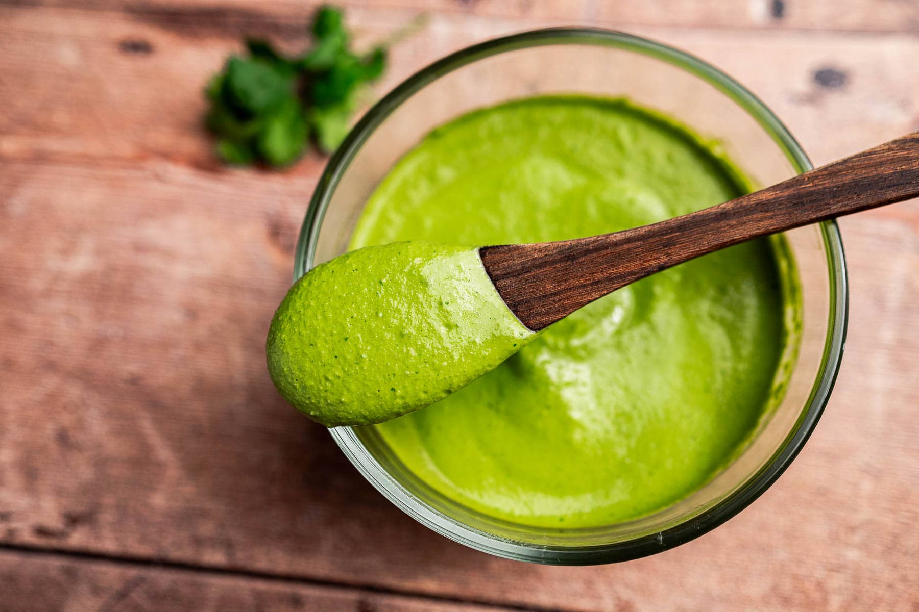 A wooden spoon holds a thick green cilantro sauce over a glass bowl filled with the same sauce, placed on a wooden surface.