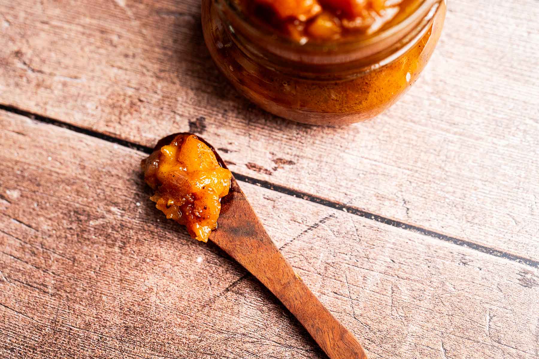 Rustic jar of peach chutney on wood table with spoonful nearby.