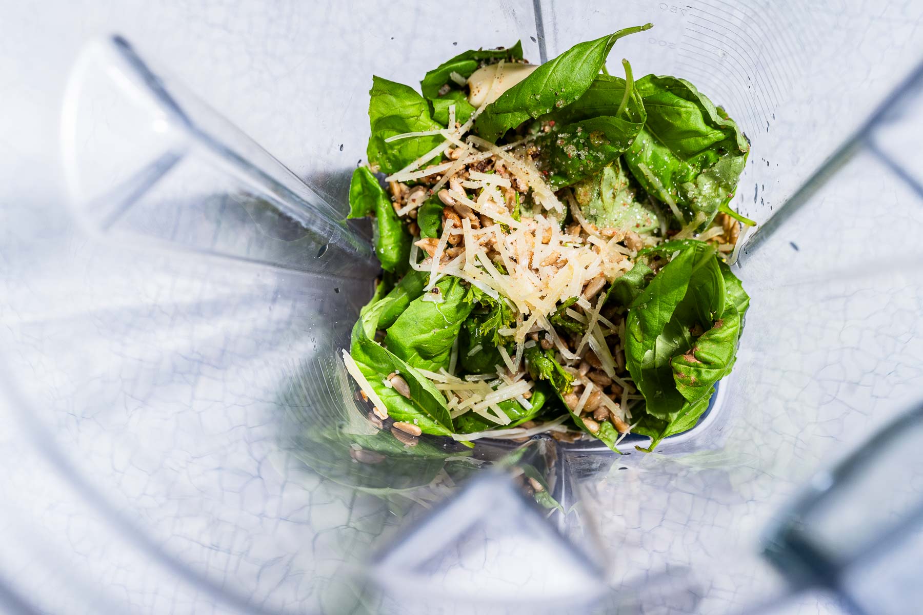 Overhead view of fresh basil leaves, grated cheese, nuts, and garlic in a blender, ready to be mixed for a delicious pesto salad dressing or sauce.