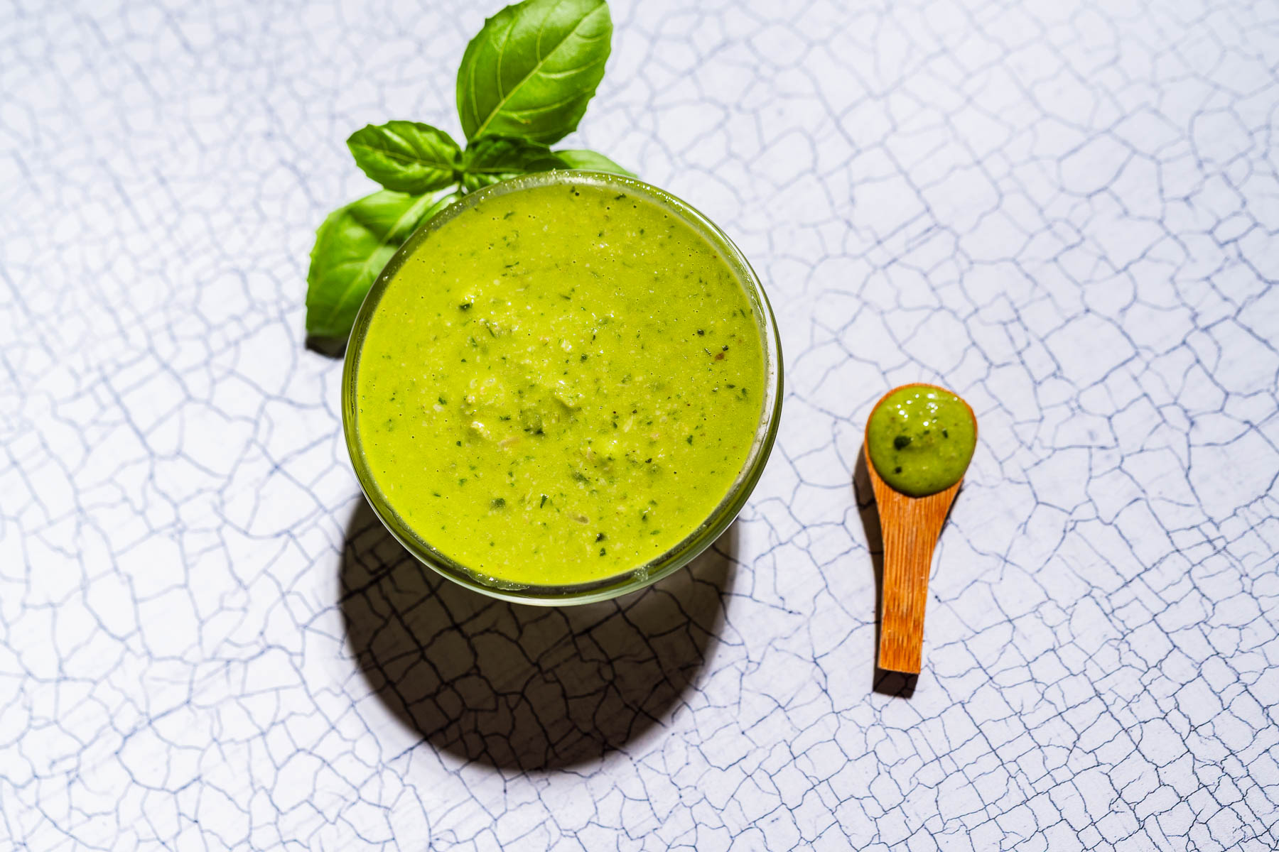 A glass bowl of green pesto salad dressing or dip, garnished with basil leaves, with a small wooden spoon holding a sample, on a cracked white surface.