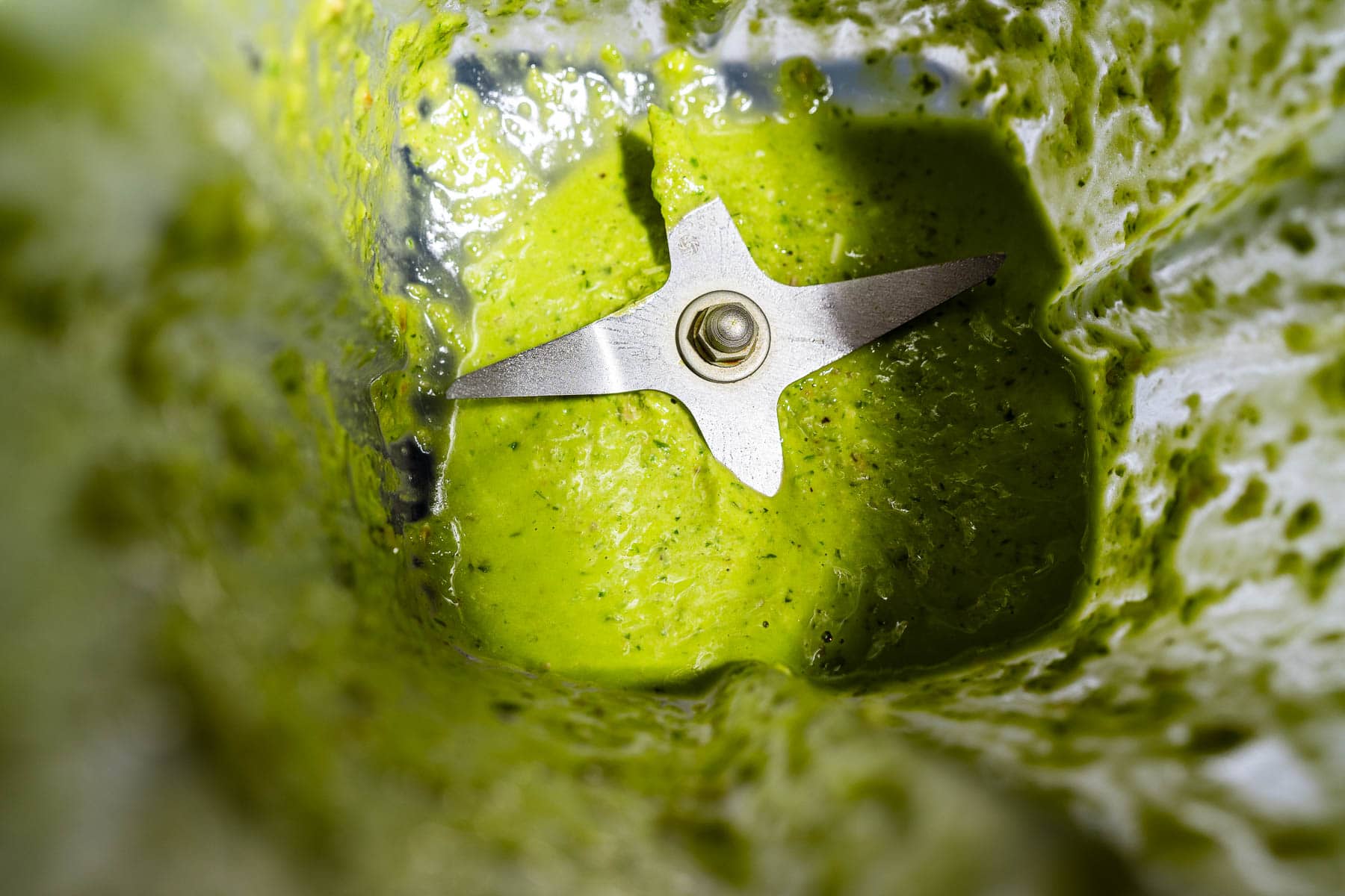 Close-up view of a blender with a vibrant green pesto salad dressing mixture and metal blades visible at the bottom.