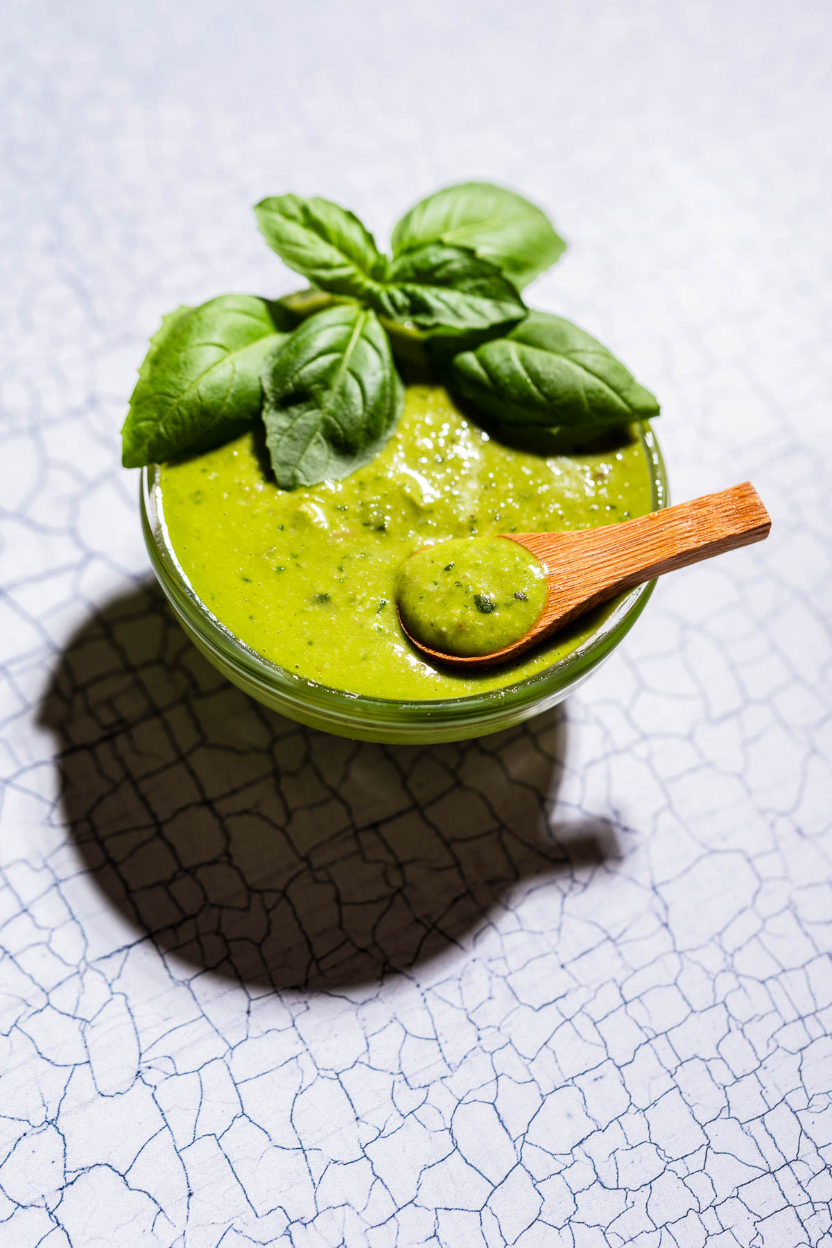 A small glass bowl of green pesto salad dressing topped with fresh basil leaves, with a wooden spoon resting in the bowl, on a white surface.