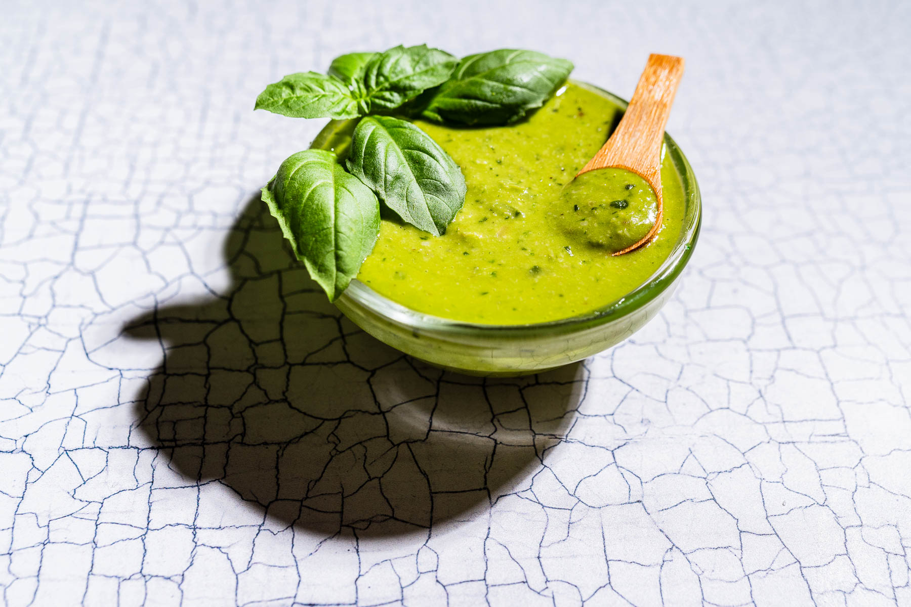 A glass bowl of green pesto salad dressing garnished with fresh basil leaves and a small wooden spoon, placed on a cracked white surface.