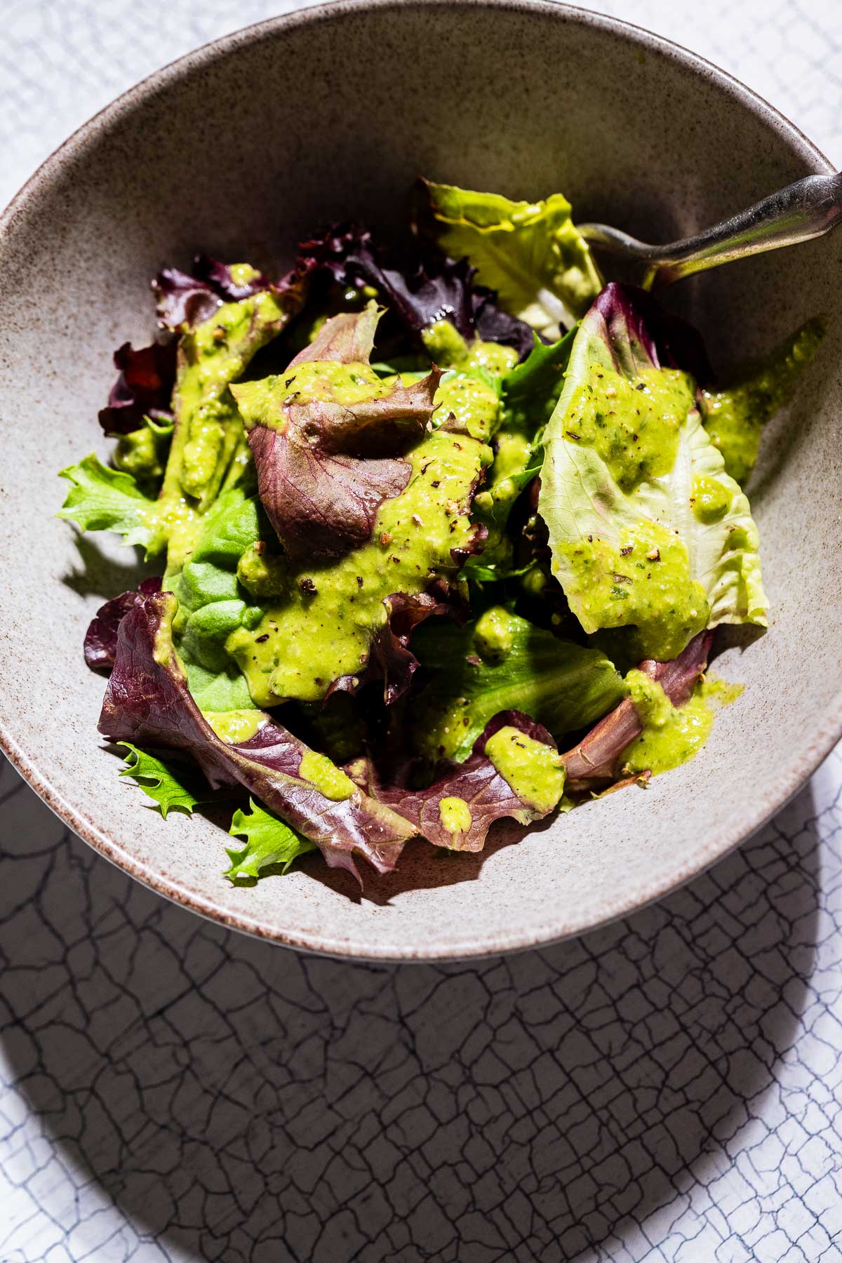 A bowl of mixed leafy greens topped with a vibrant pesto salad dressing, with a fork resting in the bowl.