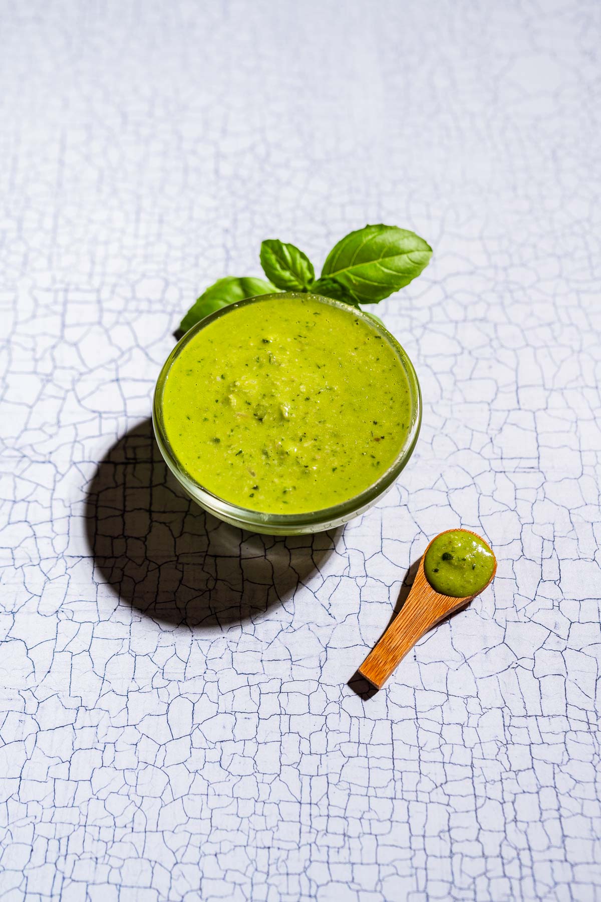 A bowl of green pesto salad dressing garnished with basil leaves sits beside a small wooden spoon filled with sauce, arranged on a white textured surface.
