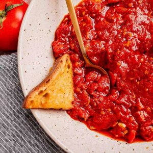 A plate of chunky matbucha tomato sauce with a gold spoon and a piece of toasted bread on the side, placed on a striped cloth next to a fresh tomato.