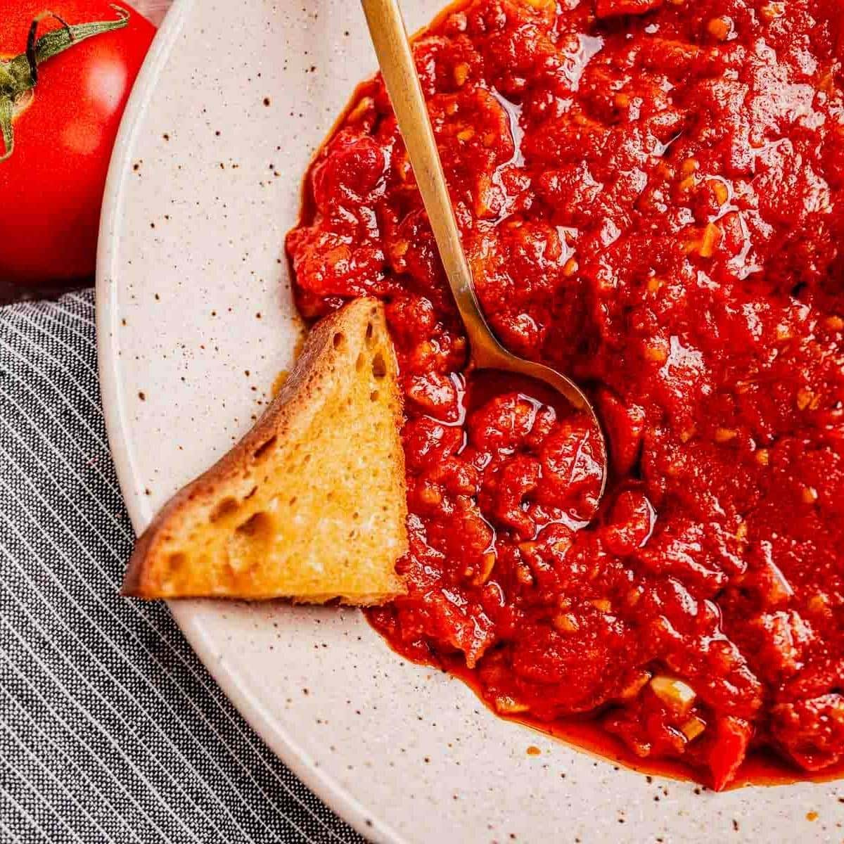 A plate of chunky matbucha tomato sauce with a gold spoon and a piece of toasted bread on the side, placed on a striped cloth next to a fresh tomato.