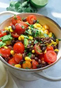 A bowl of salad containing cherry tomatoes, black beans, corn, avocado, and chopped cilantro on a light surface.