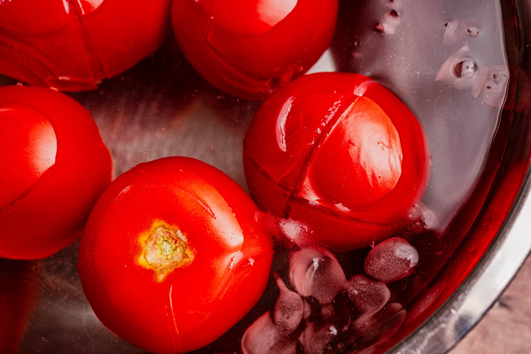 Several red tomatoes with split skins are submerged in ice water along with ice cubes, likely being peeled in preparation for making matbucha.