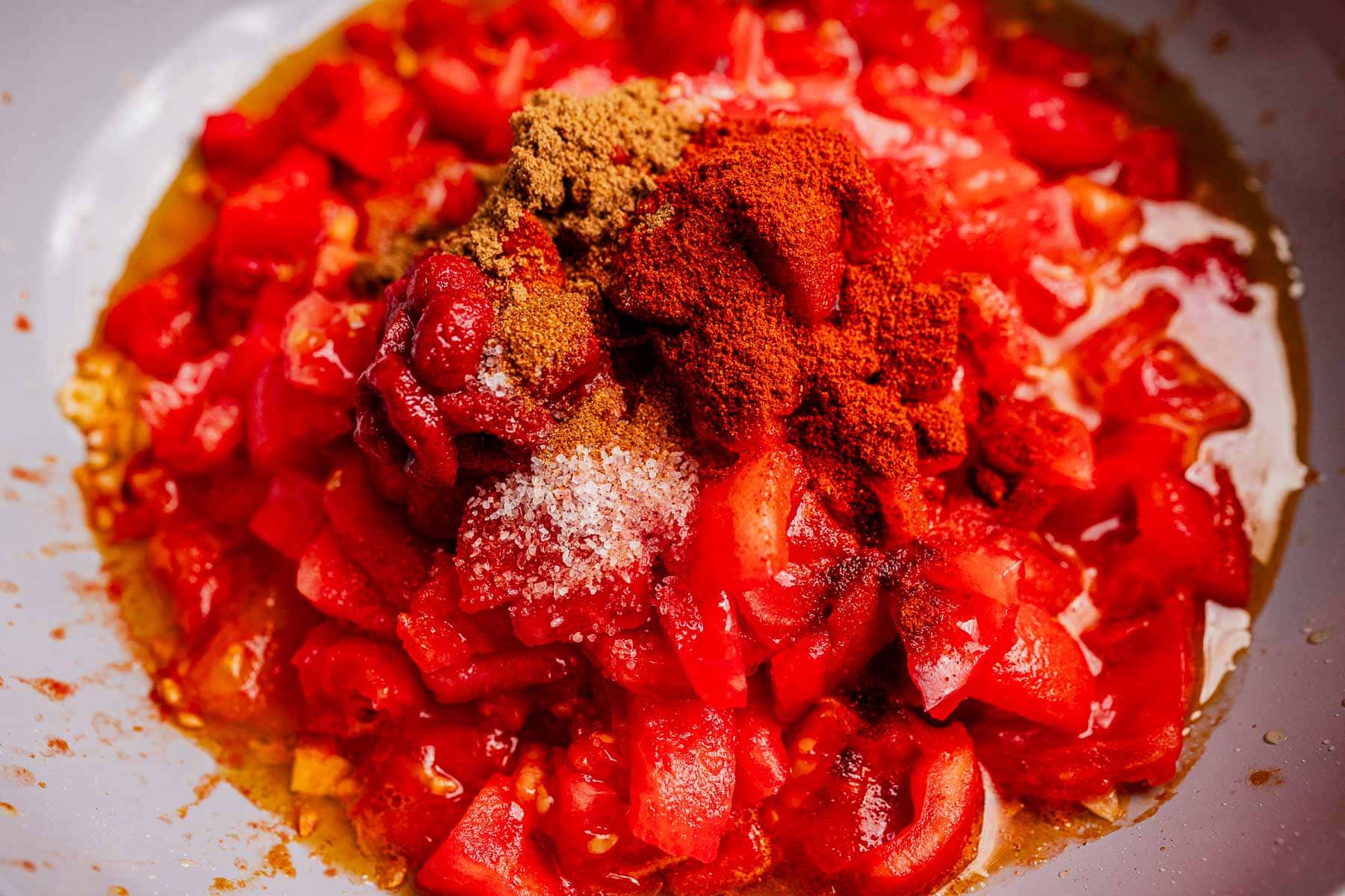 Chopped tomatoes in a bowl topped with paprika, ground spices, and salt, ready to be mixed into a flavorful matbucha.