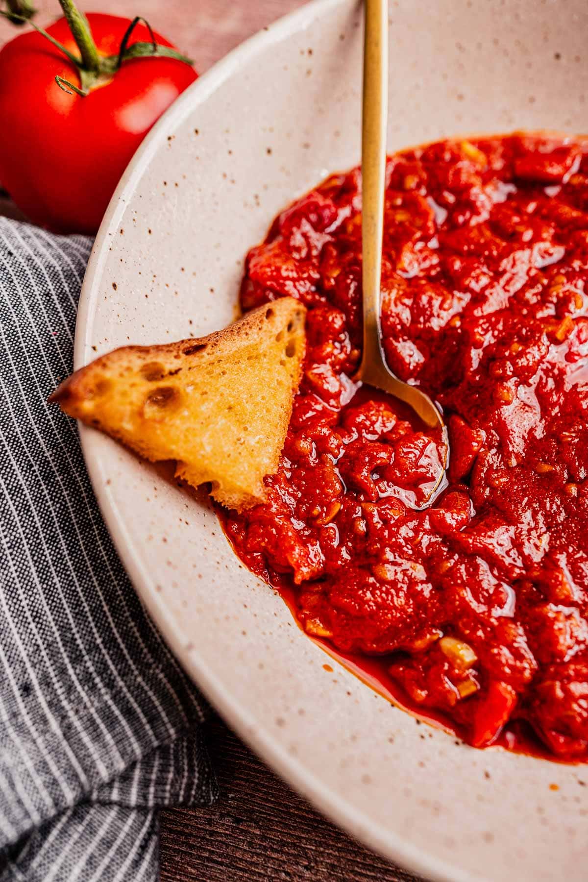 A bowl of matbucha, a rich tomato-based sauce, with a gold spoon and toasted bread sits next to a fresh tomato and striped cloth.