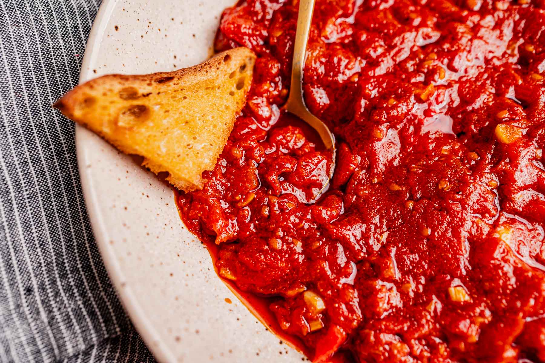 Close-up of a plate with chunky matbucha tomato sauce, a piece of toasted bread, and a gold spoon on a gray striped cloth background.