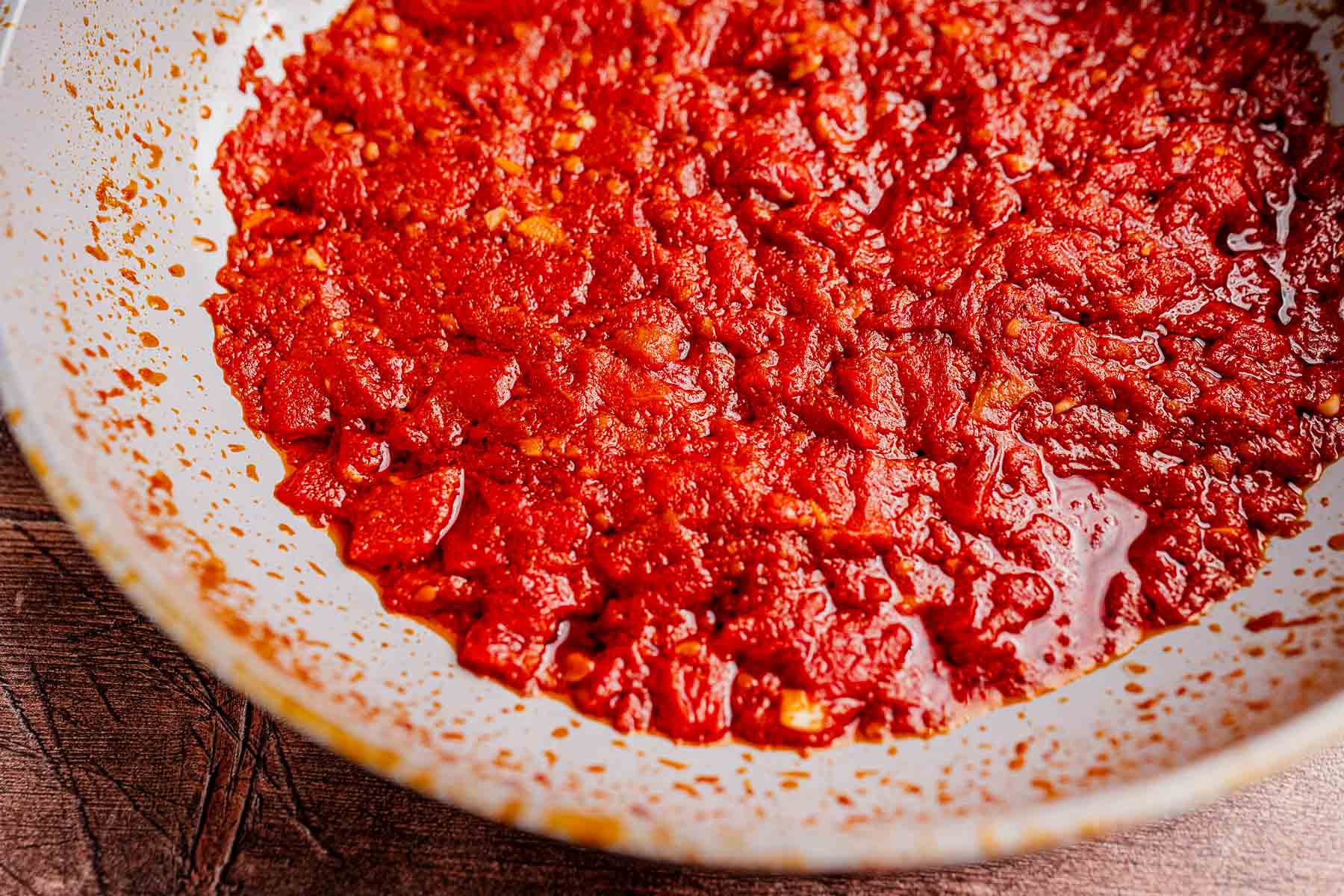 A close-up view of crushed tomatoes with juices, reminiscent of matbucha, in a white, speckled bowl on a wooden surface.