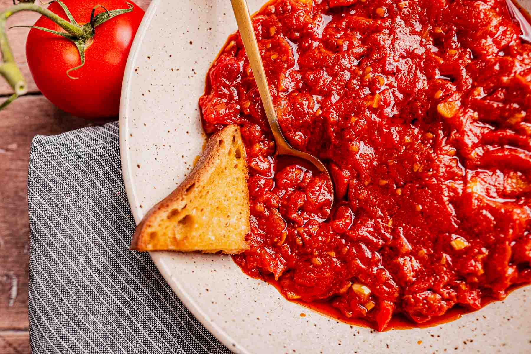 A plate of chunky matbucha tomato sauce with a piece of toasted bread and a gold fork; a fresh tomato sits nearby on a gray cloth.