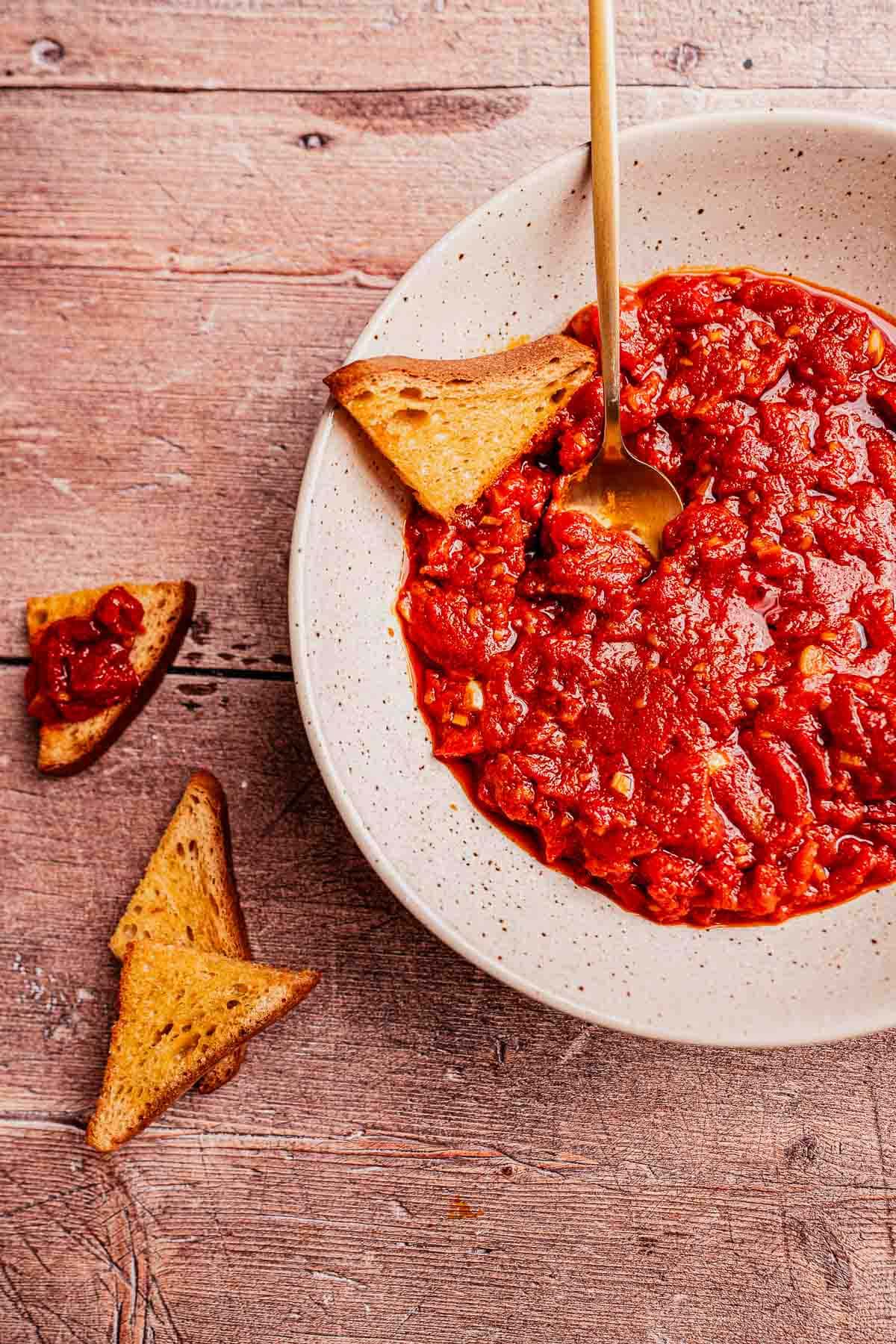 A bowl of chunky matbucha tomato dip with a spoon, served alongside toasted bread triangles on a wooden surface.