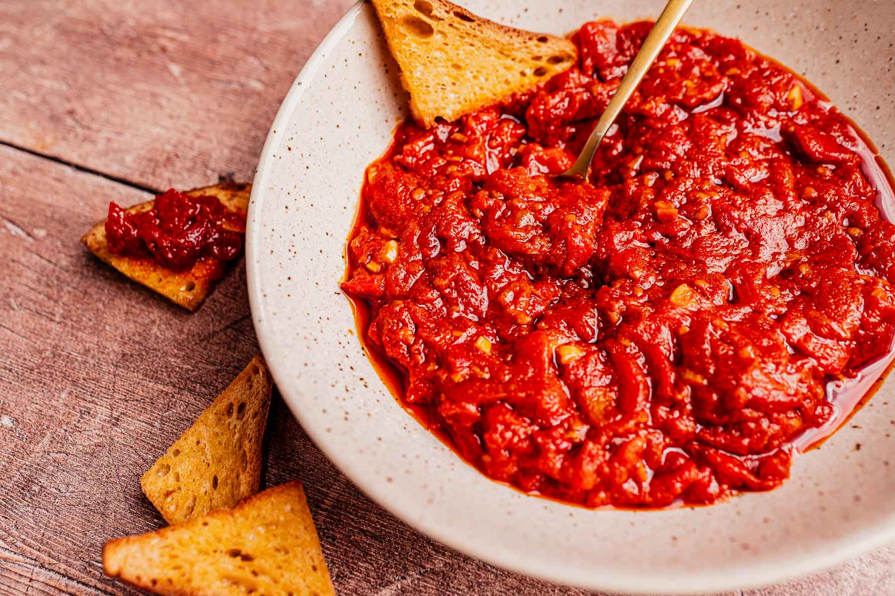 A bowl of chunky red matbucha dip with a spoon, surrounded by triangular crackers on a wooden surface.