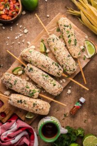 Five ears of Mexican street corn (elote) on wooden skewers are topped with cheese and herbs, served on a wooden board with lime wedges, hot sauce, and a cup of drink nearby.