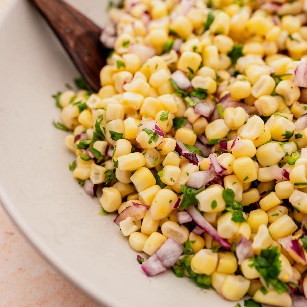 A close-up of corn salad with chopped red onions and cilantro served in a white bowl, with a wooden spoon partially visible.