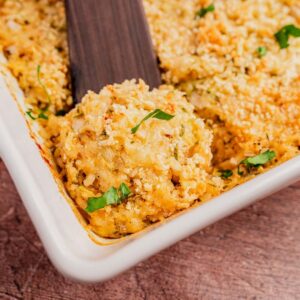 A serving of zucchini rice casserole with a golden breadcrumb topping is being scooped from a white baking dish; garnished with chopped herbs.
