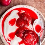 A plate with dollops of whipped cream and stewed plums in red syrup, with a spoon resting on the side. The rich, sweet stewed plums pair perfectly with the airy whipped cream for a delightful dessert.