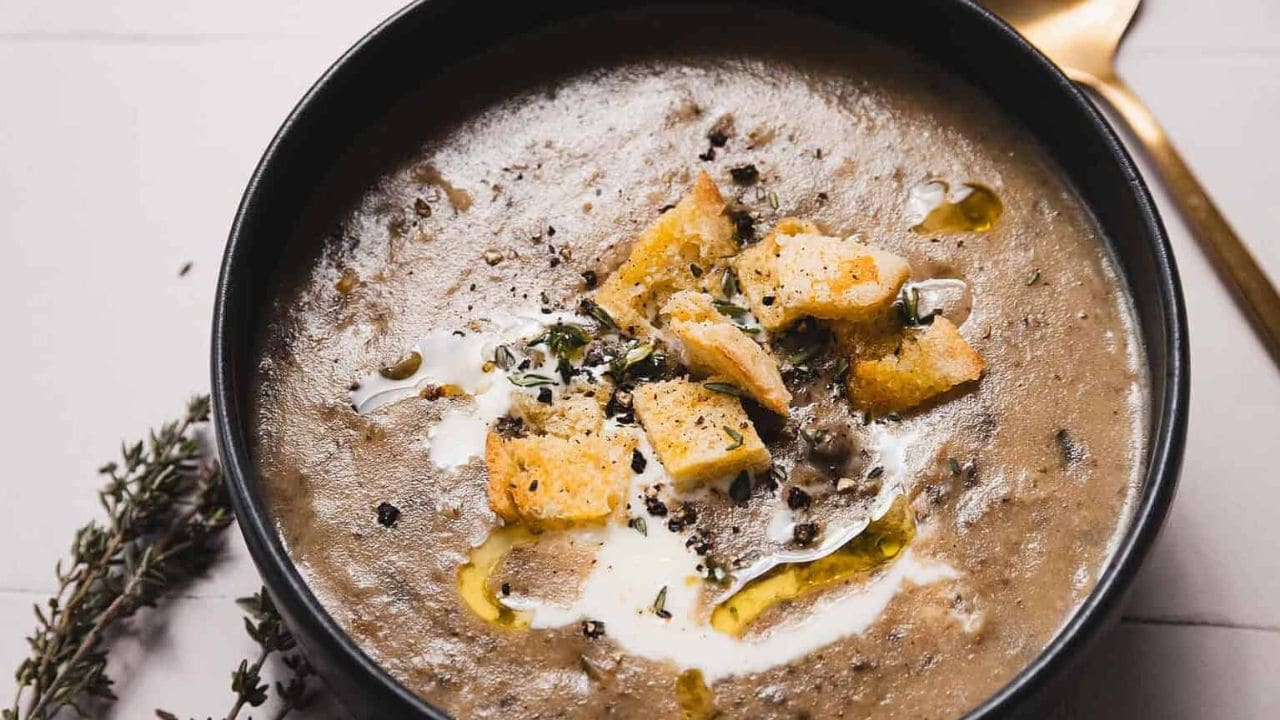 A bowl of creamy mushroom soup garnished with croutons, a drizzle of olive oil, and fresh herbs. A golden spoon lies nearby, and sprigs of thyme are placed on the table beside the bowl.