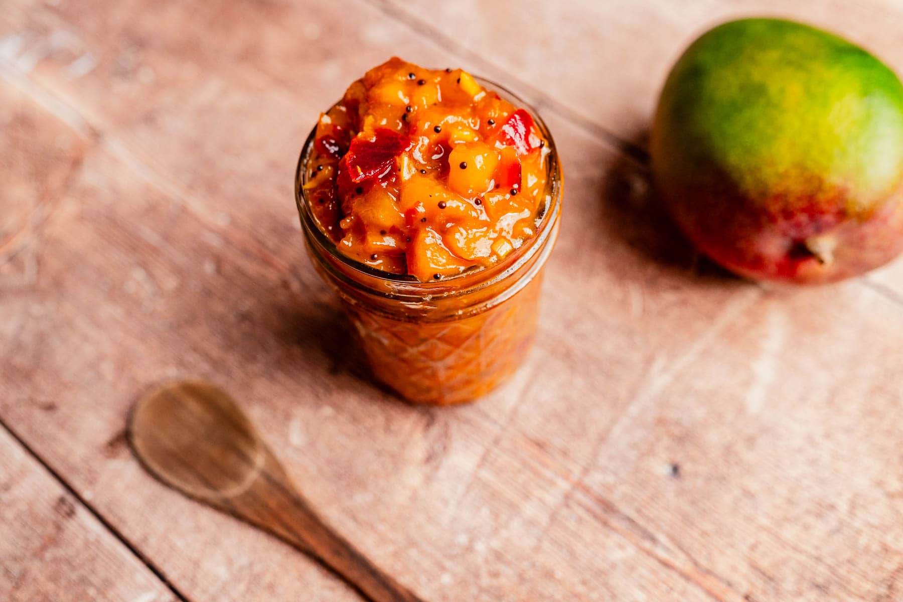 A glass jar filled with chunky mango chutney sits on a wooden surface next to a wooden spoon and a whole mango, highlighting the freshness of homemade mango chutney.