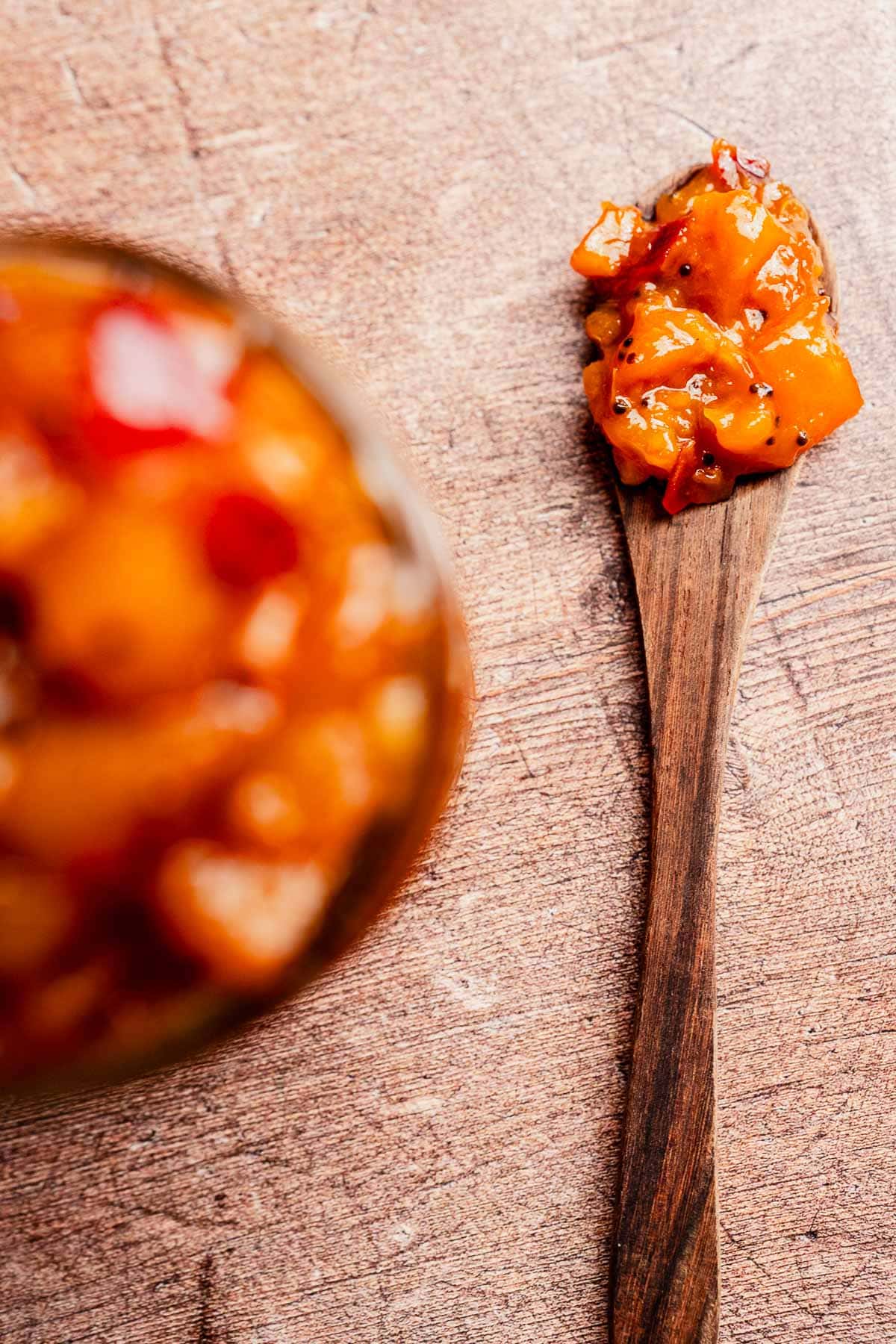 A wooden spoon with a serving of chunky orange and red mango chutney rests on a textured wooden surface, with more chutney visible in a blurred container to the left.
