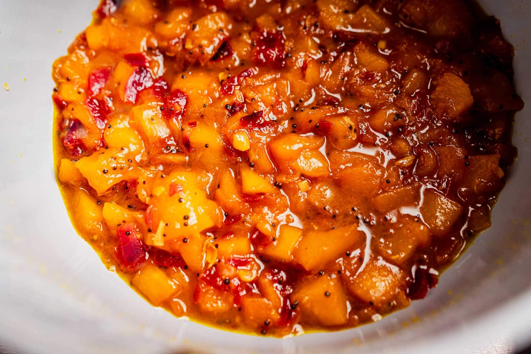 A close-up of chunky mango chutney with visible pieces of fruit and red peppers in a white bowl, showcasing its vibrant orange color.