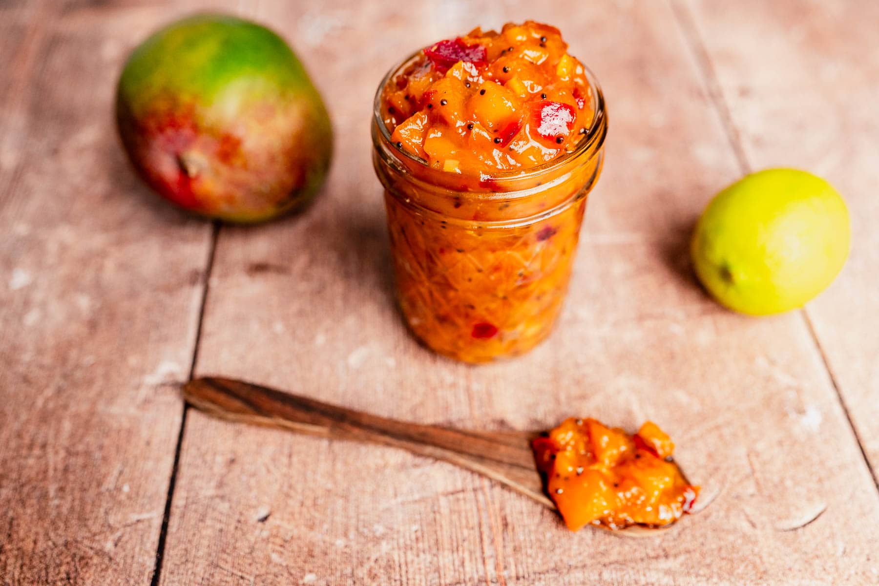A glass jar filled with mango chutney sits on a wooden surface next to a fresh mango, a lime, and a spoon topped with vibrant mango chutney.
