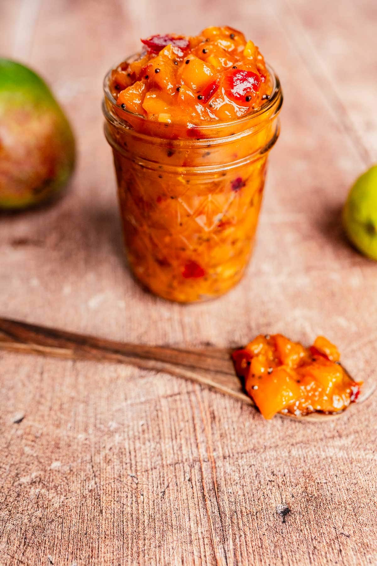 A glass jar filled with chunky mango chutney sits on a wooden surface, with a wooden spoon holding a portion of the sweet and tangy mango chutney in front.
