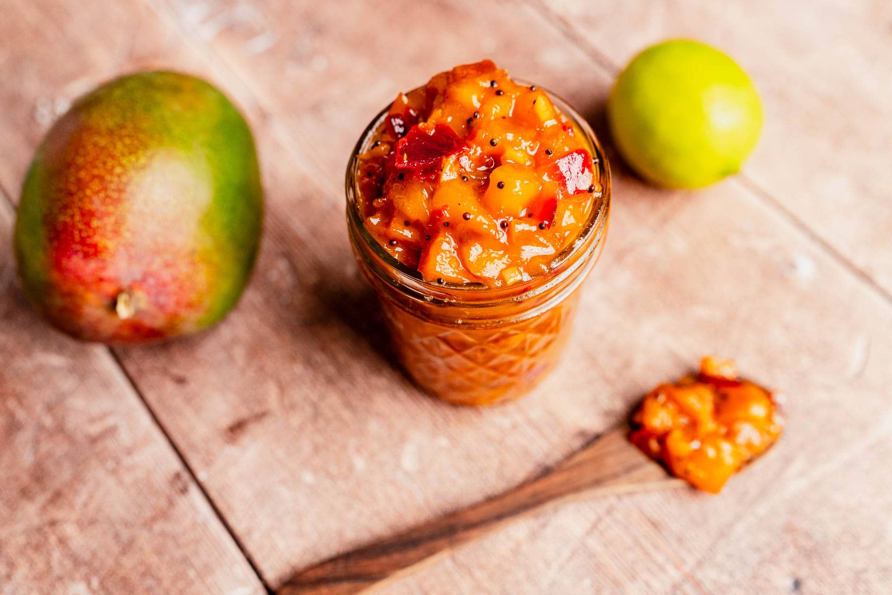 A glass jar filled with mango chutney sits on a wooden table, accompanied by a whole mango, a lime, and a spoonful of the tangy mango chutney beside it.