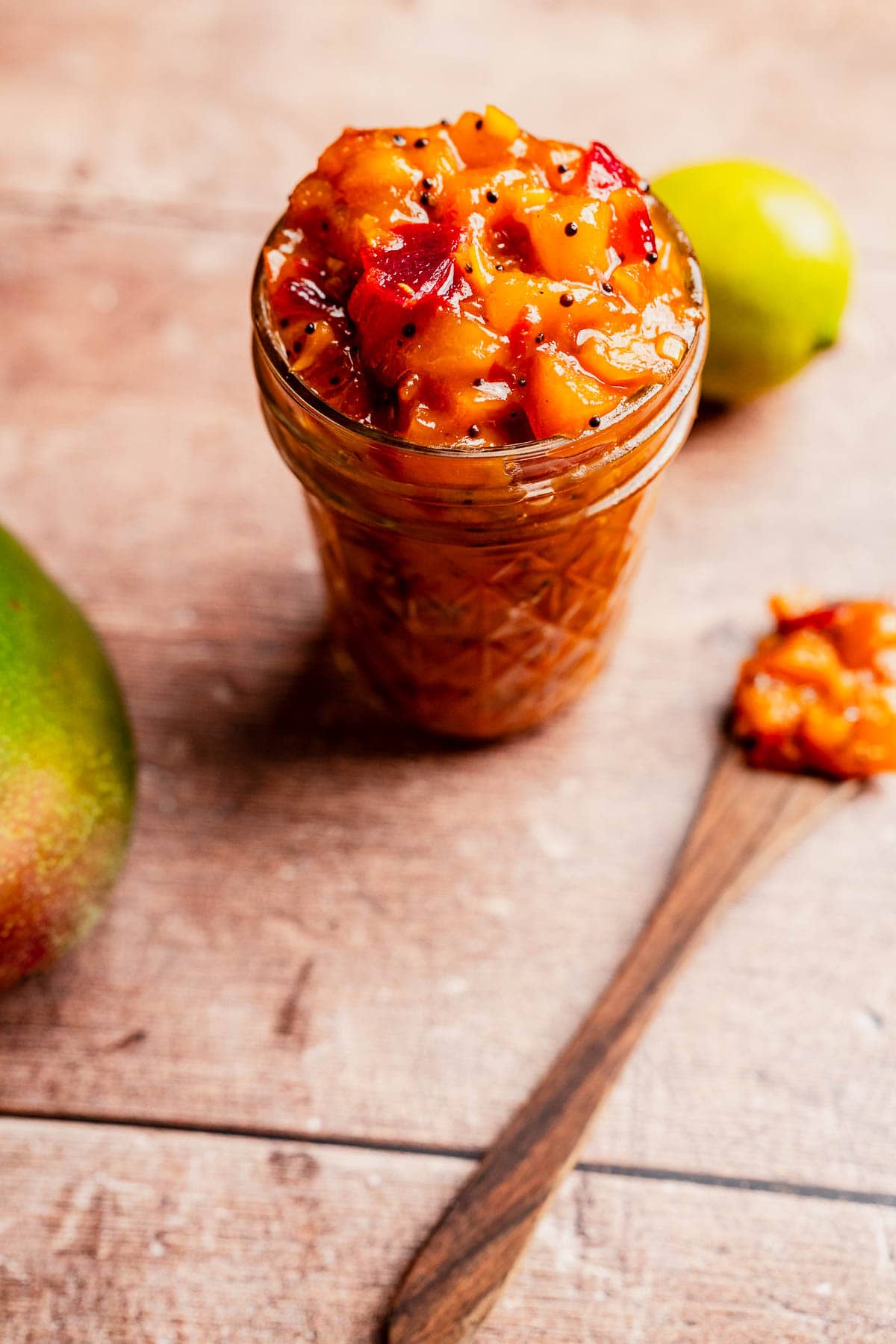 A glass jar filled with chunky mango chutney sits on a wooden surface next to a wooden spoon holding vibrant mango chutney, alongside a fresh mango and a zesty lime.