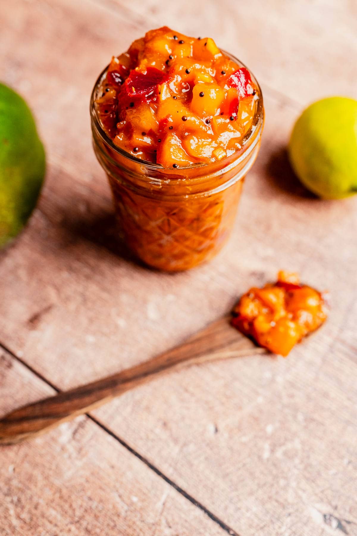 A glass jar filled with chunky mango chutney sits on a wooden surface, with a spoonful of the tangy mango chutney nearby and two whole fruits in the background.