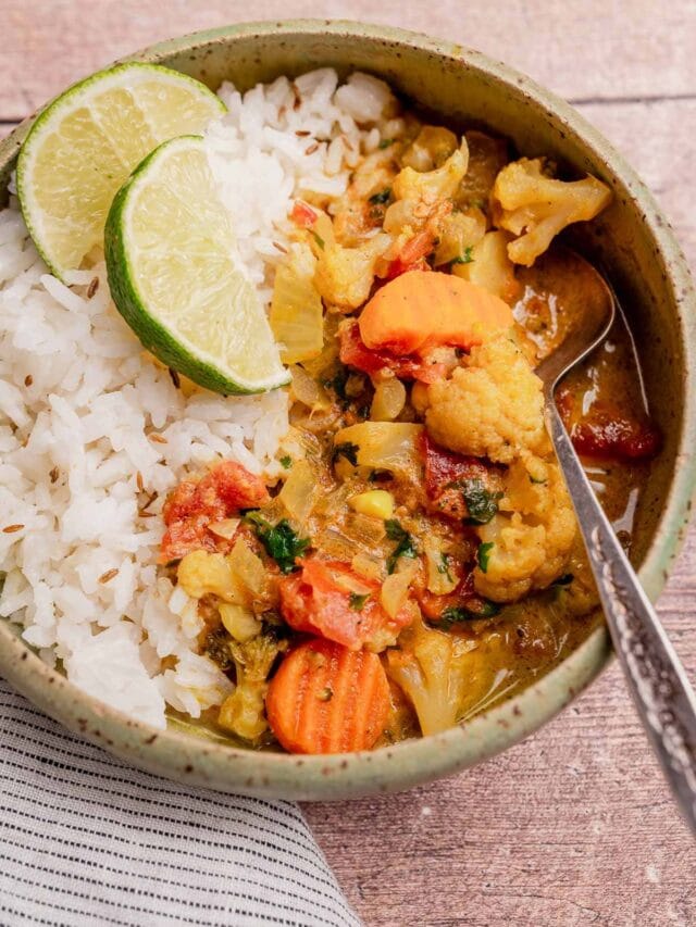 A bowl of white rice and slow cooker vegetable curry with carrots, cauliflower, and tomatoes, garnished with two lime slices and served with a spoon.