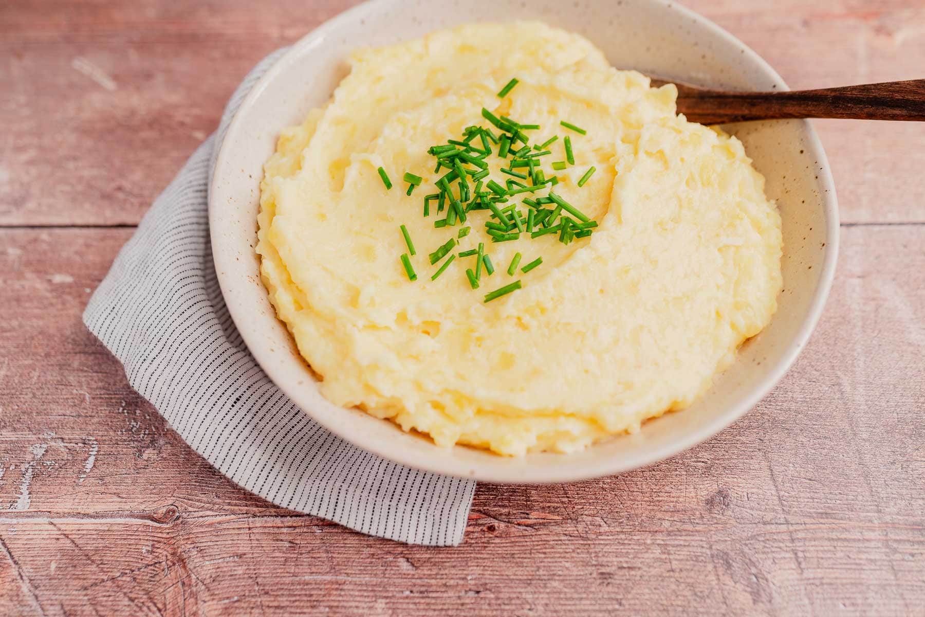 A bowl of cheesy mashed potatoes topped with chopped chives, served with a wooden spoon on a gray striped cloth.
