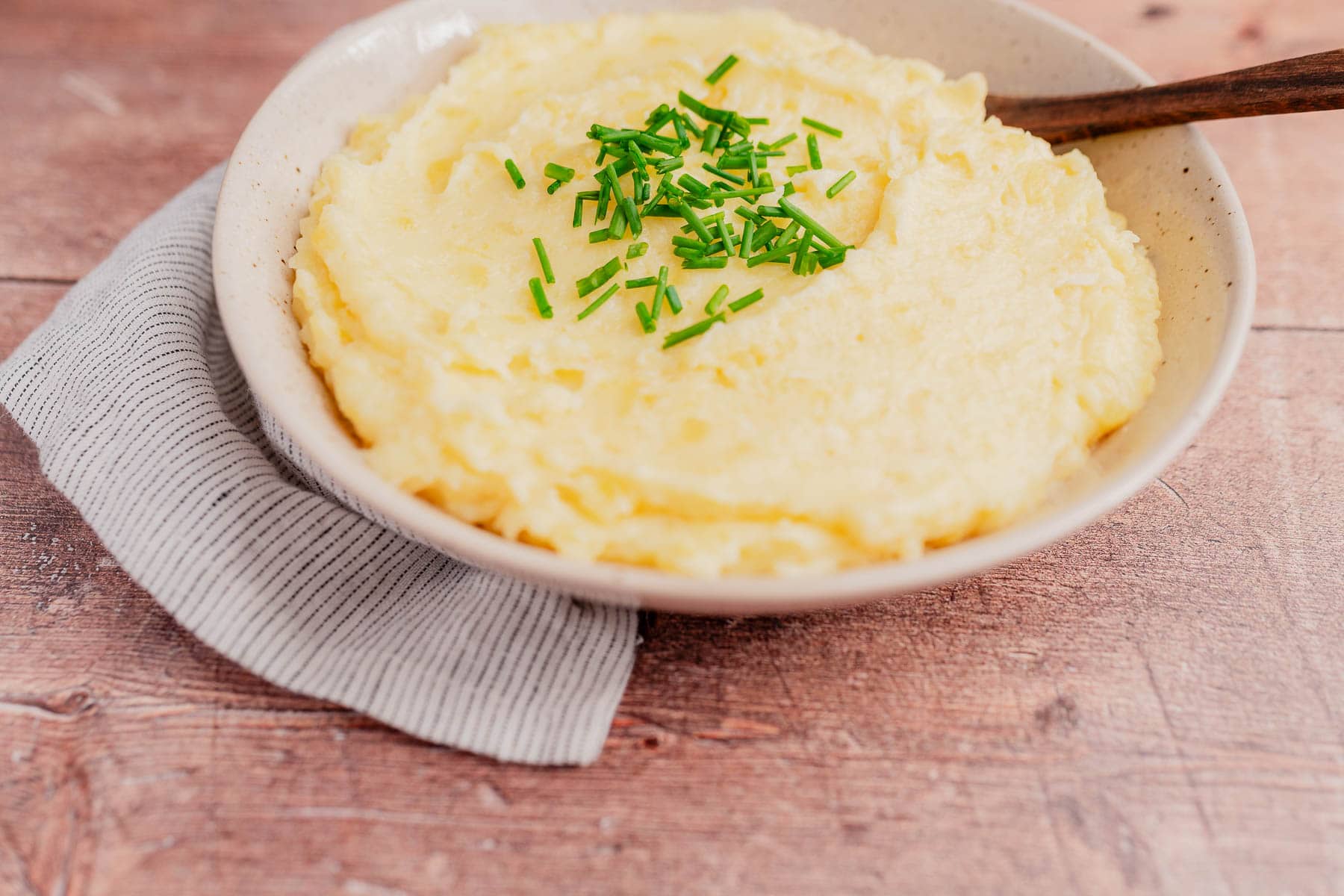 A bowl of cheesy mashed potatoes topped with chopped chives, placed on a wooden surface with a striped napkin underneath.