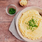 A bowl of cheesy mashed potatoes topped with chopped chives, with a wooden spoon, sits next to bowls of shredded cheese and extra chives on a wooden surface.