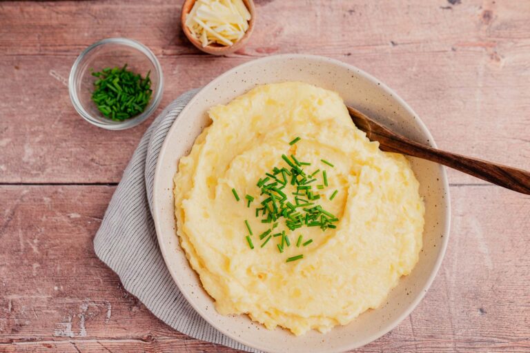 A bowl of cheesy mashed potatoes topped with chopped chives, with a wooden spoon, sits next to bowls of shredded cheese and extra chives on a wooden surface.