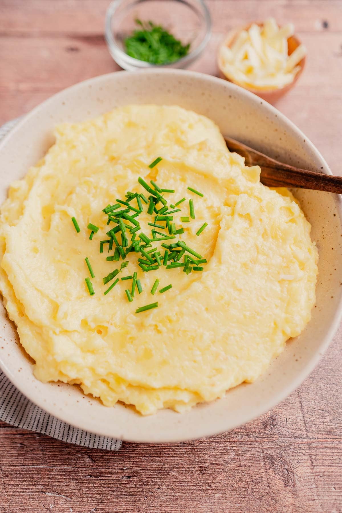 A bowl of creamy cheesy mashed potatoes topped with chopped chives, with a spoon and small bowls of shredded cheese and extra chives in the background.