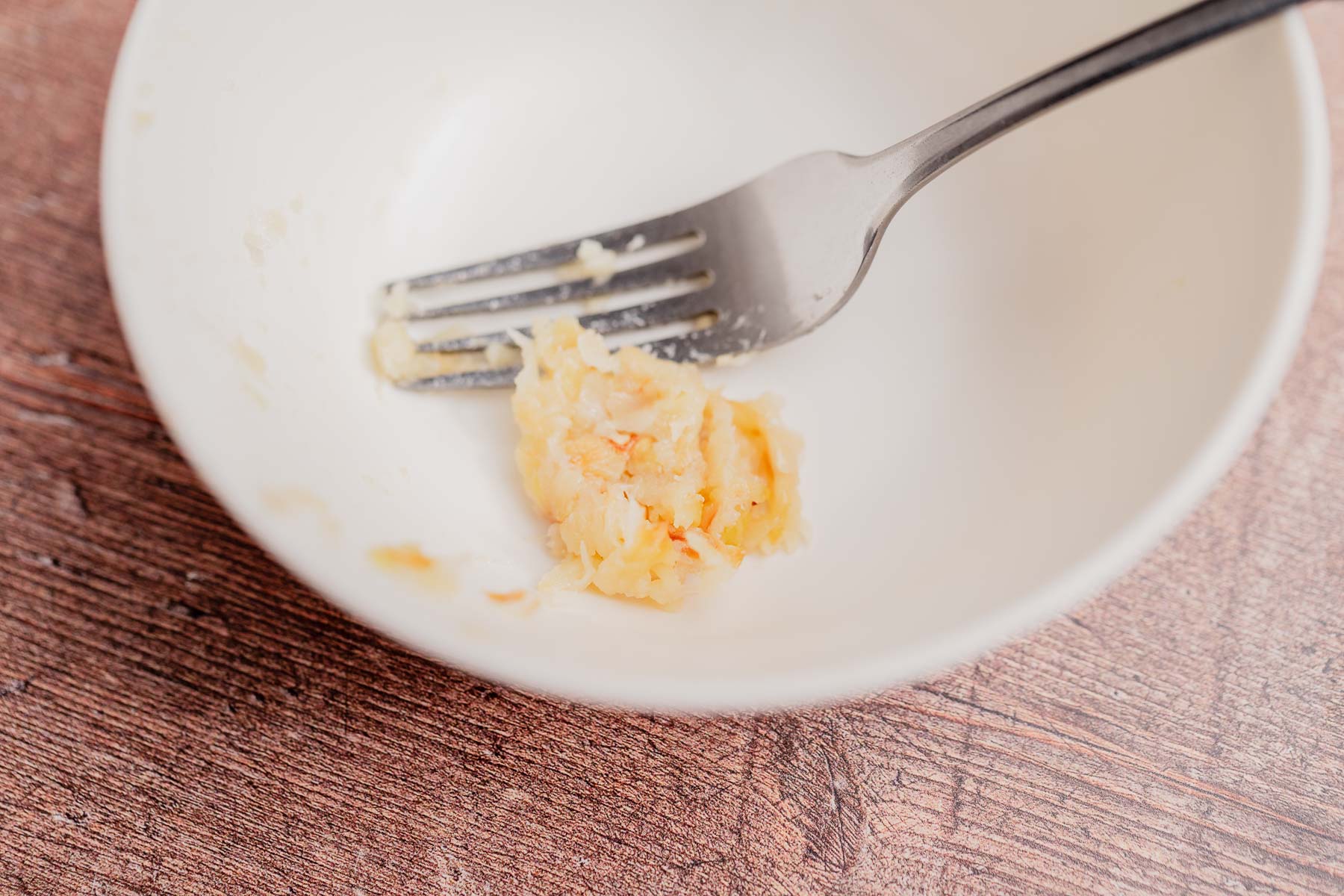 A metal fork rests in a white bowl with a small amount of cheesy mashed potatoes on a wooden surface.