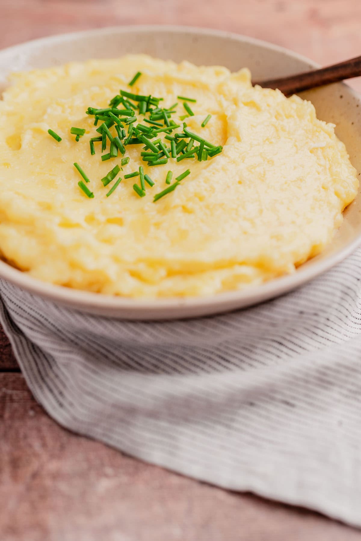 A bowl of cheesy mashed potatoes topped with chopped chives, placed on a striped cloth napkin with a spoon.