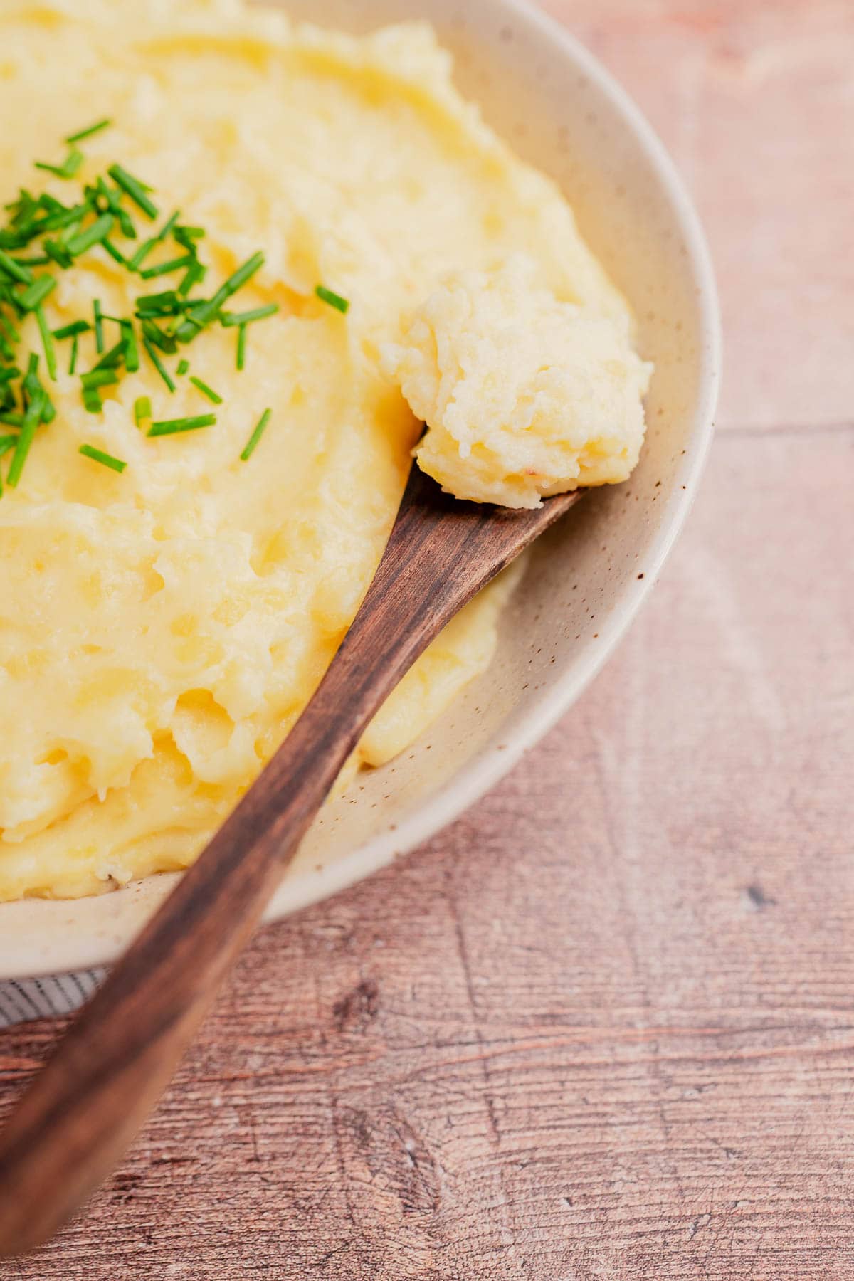 A bowl of cheesy mashed potatoes topped with chopped chives, with a wooden spoon resting on the edge holding a spoonful.