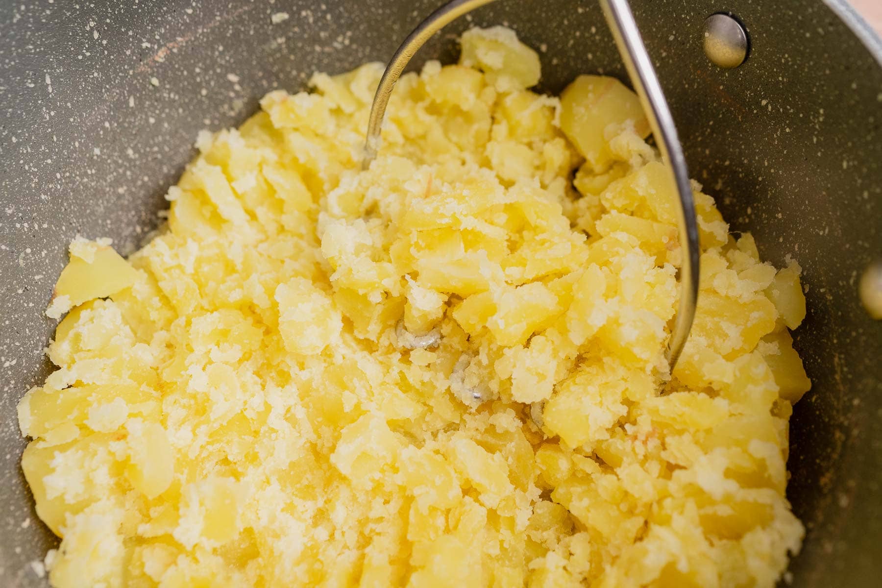 A potato masher pressing down on partially mashed boiled potatoes in a large pot, perfect for making cheesy mashed potatoes.