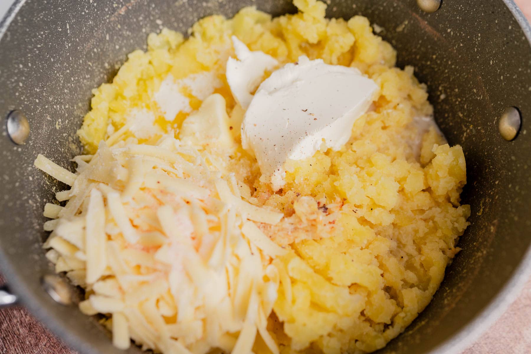 A pot containing cheesy mashed potatoes with shredded cheese and a dollop of cream cheese, ready to be mixed together.
