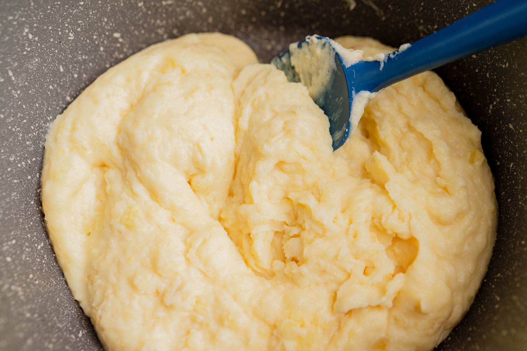 A close-up of cheesy mashed potatoes being stirred with a blue spatula in a speckled mixing bowl.